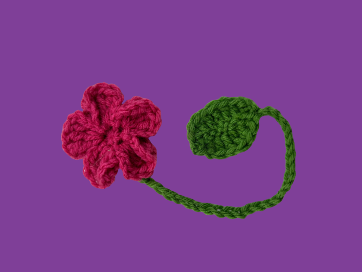 A purple background with a crocheted five-petaled pink flower with a green vine like stem and leaf
