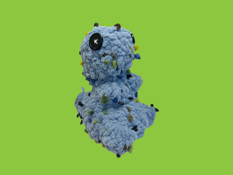 A green background with a small blue and speckled crocheted t-rex with button eyes.