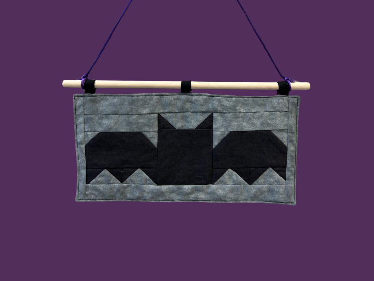 A purple background with a gray and black retangular fabric banner that depicts the shape of a bat using triangles and squares.