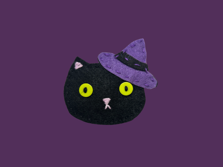 A purple background with a black cat head, made of felt, with green button eyes and a purple witch hat.