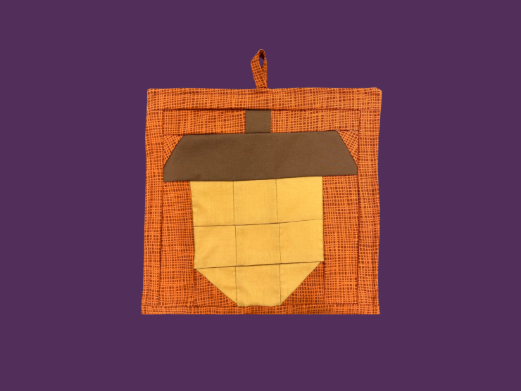 A purple backround with an orange and brown square fabric trivet with an acorn made of squares and triangles in the center
